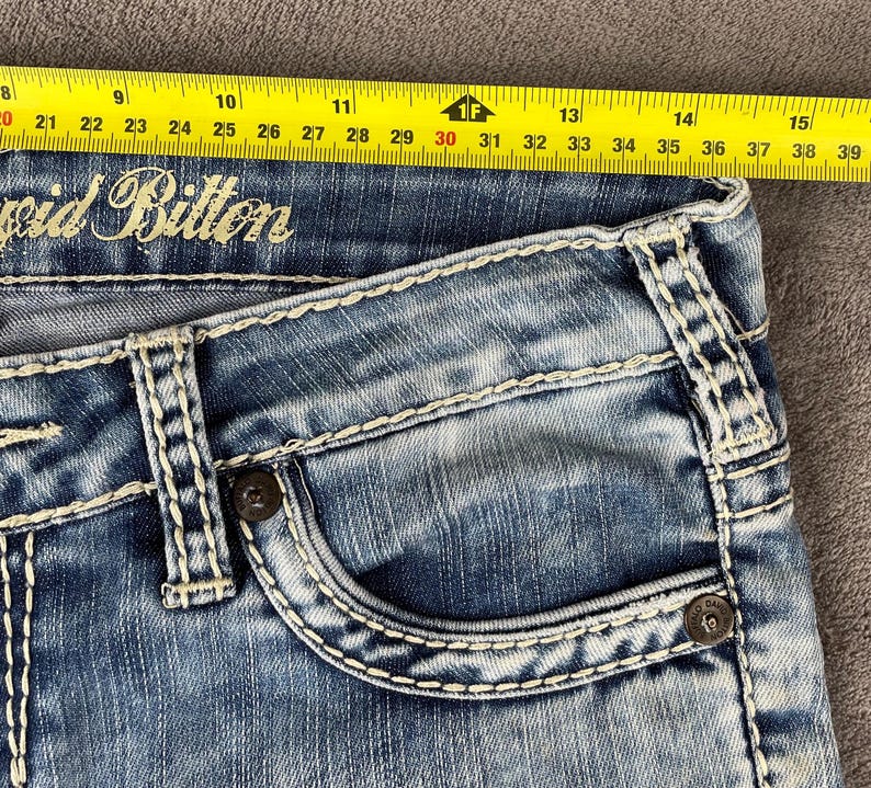 May include: Close-up of blue denim jeans with white stitching and a pocket. The jeans have the brand name "void Bitton" embroidered on them. A yellow measuring tape is placed across the top of the jeans, showing measurements from 20 to 39 inches.