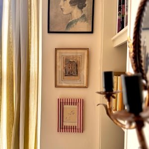 May include: Three framed pictures are displayed on a wall. The top one is a portrait of a woman in profile. Below it is a framed architectural print. The bottom frame has a red and white striped border and contains a sketch.