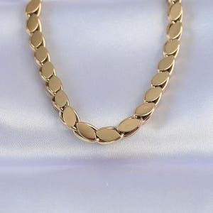 May include: A gold-colored necklace with a repeating oval link design. The necklace is displayed on a white, satin-like surface, creating a contrast that highlights the jewelry's details. The chain is made of small, flat, oval-shaped links.