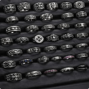 Könnte beinhalten: Eine Sammlung von Silberringen mit verschiedenen Designs, darunter Blumen-, Kreuz- und geometrische Muster. Die Ringe werden auf einer schwarzen Oberfläche präsentiert, die ihre komplizierten Details und Texturen hervorhebt. Die Ringe sind in verschiedenen Größen und Stilen erhältlich.