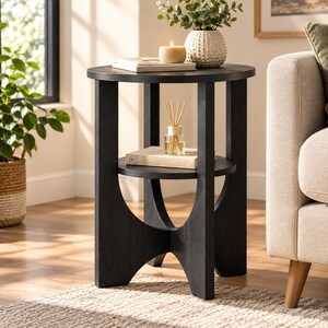 May include: A round black side table with a lower shelf, featuring three thick vertical legs that curve inward at the base. The table surface holds a white book and a yellow candle, while the lower shelf holds a glass reed diffuser and a book.