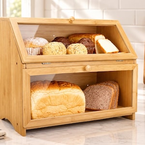 May include: A two-tier bamboo bread box with clear glass windows on the front panels. The top shelf contains assorted muffins and sliced bread, while the bottom shelf holds a loaf of white bread and several slices of dark rye bread.