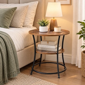 May include: A round side table with a dark wood grain finish top and a lower shelf, supported by a black metal frame. The table holds a white ribbed planter with a green plant, a wooden lamp with a white shade, and a candle. Two books are on the shelf.