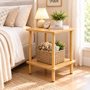 May include: A square light-toned wooden side table with four cylindrical legs and a lower shelf. The top surface holds a pleated lamp, a round analog clock, a potted green plant, and a book. A woven basket containing a teddy bear sits below.