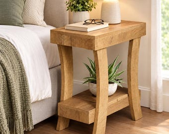 May include: A light brown wooden end table with a rectangular top and a lower shelf. The table features four curved legs and a wood grain finish. It is positioned next to a bed and holds a white lamp, a book, black glasses, and a plant.