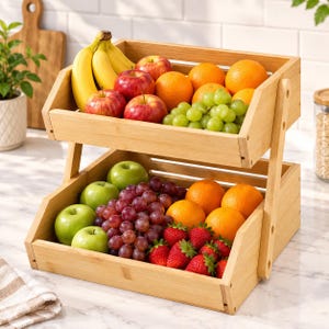 May include: A two-tier bamboo fruit basket with an open-slat design. The top tier holds yellow bananas, red apples, oranges, and green grapes. The bottom tier contains green apples, red grapes, oranges, and red strawberries in a wooden frame.