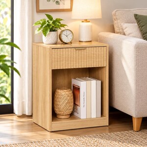May include: A light oak nightstand featuring a single top drawer with a slim gold handle and an open lower shelf. The shelf contains a woven rattan lamp and a row of books, including one titled The Kinfolk Home.