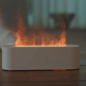 Peut inclure: Cheminée de table rectangulaire blanche avec un effet de flamme simulé. Les flammes sont orange et jaune, créant une ambiance chaleureuse. Une petite plante en pot est à droite sur une surface en bois.