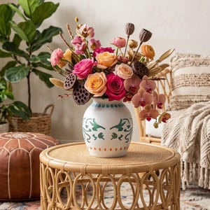 Può includere: Un vaso in ceramica bianca dal corpo arrotondato, caratterizzato da un motivo decorativo di forme a foglia verde e punti arancioni. Il vaso è riempito con una vivace composizione floreale di rose rosa e pesca, orchidee e baccelli di loto marroni.