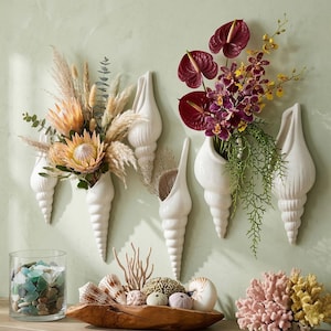 Op de afbeelding: Witte schelpvormige wandvazen gevuld met gedroogde bloemen en verse bloemen. Een glazen pot met zeeglas en een houten kom met schelpen en koraal zijn ook te zien. Het kleurenpalet omvat wit, beige, roze en bordeaux.