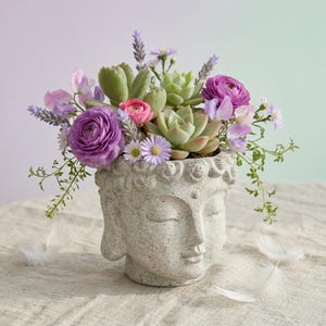 May include: A stone Buddha head planter filled with a mix of succulents and flowers. The arrangement features purple ranunculus, lavender, and pink blooms. The planter is grey and has a peaceful expression.