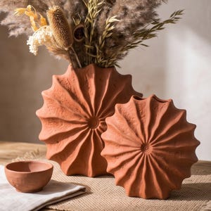 Pode incluir: Dois vasos de cor terracota com um design circular plissado em forma de raios de sol e uma superfície texturizada. Uma pequena taça de terracota a condizer encontra-se à esquerda. O vaso maior contém um arranjo de flores secas em tons de bege e castanho.