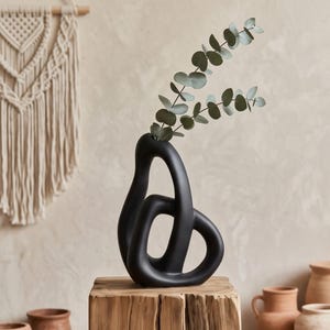 Könnte beinhalten: Eine mattschwarze Keramikvase mit einem abstrakten, hohlen und ineinander verschlungenen Schleifendesign. Die Vase hat eine glatte, geschwungene Silhouette und enthält einen Zweig mit grünen Eukalyptusblättern und kleinen, runden Blättern.