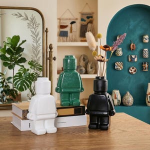 May include: Three ceramic vases shaped like a person, in white, green, and black. The black vase contains dried flowers. The vases are on a wooden surface, with a mirror and other decorative items in the background.