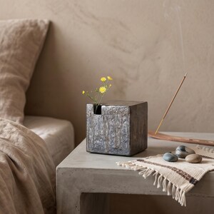 May include: A dark, metallic-toned cube-shaped ceramic vase with a textured, uneven surface. A single small stem with yellow flowers is placed in the top opening. Beside the vase, an incense stick burns on a wooden holder near three smooth stones.