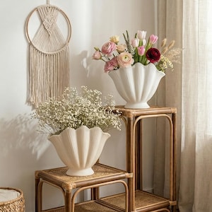 May include: Two white, shell-shaped vases filled with flowers sit on woven rattan stands. One vase holds pink and red flowers, the other holds white flowers. A macrame wall hanging is visible in the background.