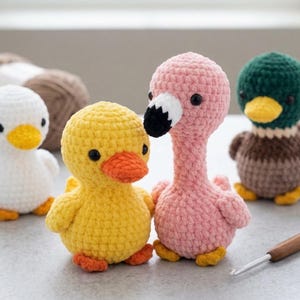 Op de afbeelding: Een verzameling handgemaakte gehaakte dierenfiguren, waaronder een wit eendje, een geel eendje, een roze flamingo en een groene en bruine eend. De figuren hebben zwarte ogen en oranje of gele snavels en voeten. Een haaknaald en knutselspullen zijn ook zichtbaar.