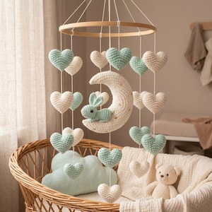 May include: A handmade baby mobile featuring a wooden ring, crocheted hearts in cream and mint green, a cream-colored moon with gold stars, and a mint green bunny. The mobile hangs above a wicker bassinet.