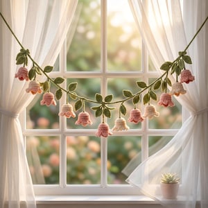 May include: A handcrafted crochet garland featuring pink and cream-colored bell-shaped flowers and green leaves. The garland is suspended in front of a window with sheer white curtains, creating a delicate and charming decorative piece.