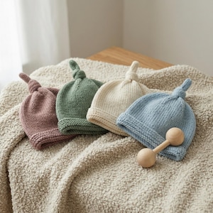 Puede incluir: Cuatro gorros de bebé de punto en tonos rosa, verde, crema y azul, cada uno con una parte superior anudada. Una sonaja de madera descansa junto al gorro azul. Los gorros se exhiben sobre una manta suave y texturizada.