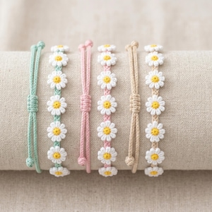 May include: Five daisy chain bracelets in pastel shades: mint green, light green, pink, beige, and tan. Each bracelet features crocheted white daisies with yellow centres. The bracelets are displayed on a neutral-coloured fabric.