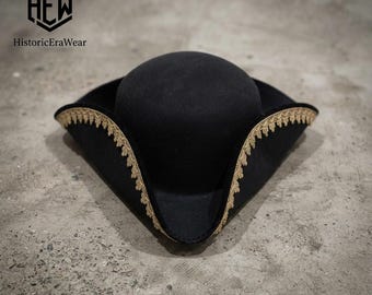 May include: A black tricorn hat featuring a rounded crown and a wide, upturned brim. The edges of the brim are adorned with a decorative gold-colored trim with a scalloped pattern. The logo for HistoricEraWear is visible in the upper left corner.
