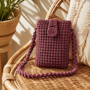 May include: A burgundy crochet phone bag with a matching strap and a button closure. The bag is rectangular with a textured stitch pattern. The strap is a braided design. The bag is sitting on a wicker chair.