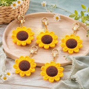 May include: Five handmade crochet sunflower keychains. Each keychain features a yellow sunflower with a brown center and a gold-colored clasp. The keychains are arranged on a wooden tray, with a background of daisies and greenery.