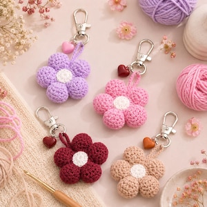 May include: Four handmade crochet flower keychains in shades of purple, pink, burgundy, and beige. Each flower has a white center and a heart charm. Silver-tone metal clasps are attached.
