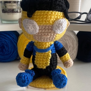 Op de afbeelding: Een handgemaakte gehaakte pop van een superheldenpersonage, met een geel en blauw kostuum met zwarte accenten. De pop heeft witte oogvormige details, een bruin gezicht en zwart haar. Hij zit op een heldere, ronde basis.