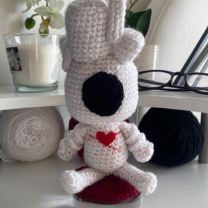 Peut inclure: Une poupée crochetée blanche avec un grand œil circulaire noir, un écusson en forme de cœur rouge sur la poitrine avec des détails cousus en rouge, et un nœud blanc sur le dessus de la tête. La poupée présente des mailles visibles et un corps arrondi.