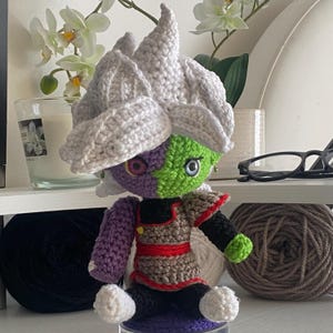 Op de afbeelding: Een handgemaakte gehaakte pop met een groot, stekelig wit kapsel, een tweekleurig gezicht met een paarse en een groene kant, en een gedetailleerde outfit in bruin, zwart, rood en groen. De figuur heeft opvallende ogen en kleine oorbellen.