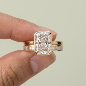 Op de afbeelding: Een gouden ringenset met een grote rechthoekige radiant-geslepen middensteen in een zetting, gecombineerd met een bijpassende ring die is ingelegd met een rij kleine diamantjes in baguette-slijpvorm. Het metaal is gepolijst geelgoud.