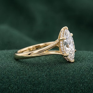 Op de afbeelding: Een gouden ring met een grote marquise-geslepen middensteen omringd door een halo van kleine ronde steentjes. De ringband heeft een gespleten ontwerp dat aansluit op de zetting, die boven de band uitsteekt om de gefacetteerde steen te tonen.