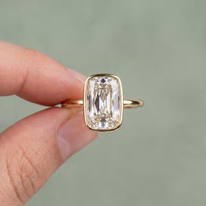 Könnte beinhalten: Ein Goldring mit einem großen, rechteckigen, im Kissenschliff facettierten, klaren Edelstein, der in einer glatten, polierten Goldzargenfassung sitzt. Die Ringschiene ist dünn und rund und aus demselben Gelbgold wie die Fassung gefertigt.