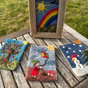 Op de afbeelding: Een houten, ingelijst viltkunstwerk toont een regenboog, een zon en een vogel. Drie andere viltkunstwerken tonen een boom, een meisje met een vlieger en een sneeuwpop. De kunstwerken liggen op een houten tafel.
