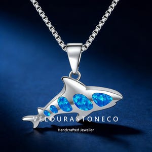 May include: A silver necklace featuring a pendant shaped like an orca whale. The whale pendant has blue opal inlays and is suspended from a silver chain. The text "VELOURASTONECO Handcrafted Jeweller" is visible.