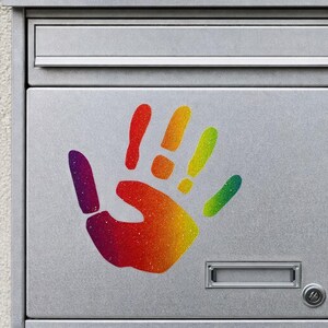 Op de afbeelding: Een zilveren brievenbus met een kleurrijk handafdrukontwerp. De handafdruk is een kleurengradient van rood, oranje, geel, groen en paars. De brievenbus heeft een gleuf voor post en een kleine deur met een slot.