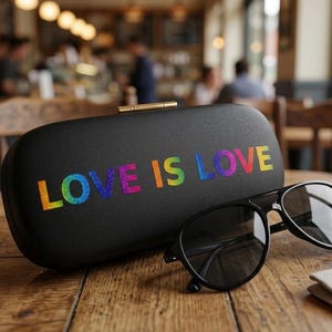 Könnte beinhalten: Ein schwarzes Brillenetui mit den Worten "LOVE IS LOVE" in Regenbogen-Glitzerbuchstaben. Neben dem Etui befindet sich eine schwarze Sonnenbrille. Die Artikel befinden sich auf einem Holztisch in einem Café.