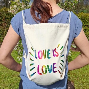 Op de afbeelding: Een crèmekleurige rugzak met trekkoord met de woorden "LOVE IS LOVE" in regenboogkleuren gedrukt. De tekst is omgeven door stralende lijnen. De tas wordt over de schouders gedragen.
