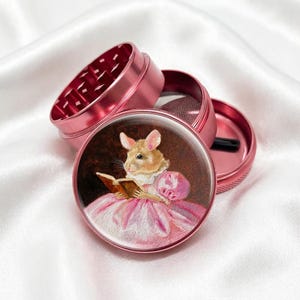 May include: A disassembled pink metal grinder with a detailed illustration of a mouse reading a book, wearing a pink dress. The grinder is composed of multiple parts, including a mesh screen and a grinding chamber.