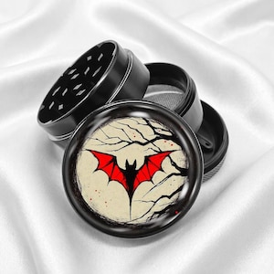 May include: Black metal grinder featuring a red bat design on the lid. The bat is set against a cream background with a tree branch design. The grinder is open, showing the grinding teeth and mesh screen.