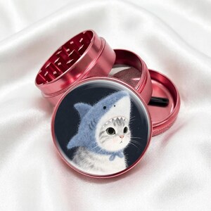 May include: A disassembled pink metal grinder with a cat wearing a shark costume on the lid. The grinder is open, revealing the internal components. The cat illustration is on a black background.