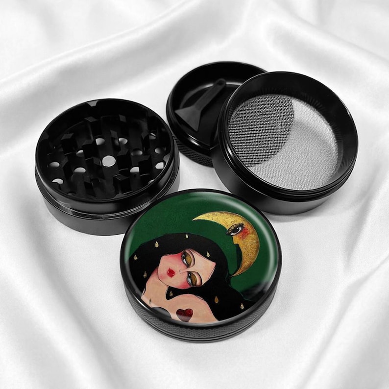 May include: Black metal herb grinder with a detailed illustration on the lid. The artwork features a woman with long black hair, a crescent moon, and a heart. The grinder is disassembled, showing its various components.
