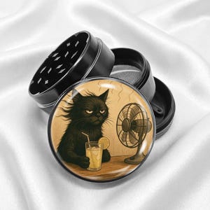 May include: Black grinder with a humorous illustration of a grumpy black cat drinking a beverage with a lemon slice, next to a fan. The grinder is black and has a diamond-shaped pattern on the top.