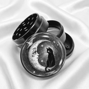 Peut inclure: Grinder noir et blanc avec un motif céleste. Le couvercle présente un croissant de lune avec un visage, des étoiles et un chat noir. Le grinder est en métal et possède plusieurs compartiments.