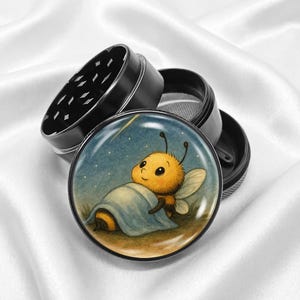 May include: Black metal herb grinder with a whimsical illustration of a cartoon bee resting under a blanket. The bee is yellow and orange with a smiling face. The grinder is open, revealing the grinding teeth and mesh screen.