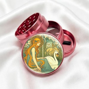May include: A disassembled pink metal grinder with a detailed illustration on the lid. The illustration depicts a woman with long orange hair and a swan in a serene, Art Nouveau style scene. The grinder is on a white satin surface.