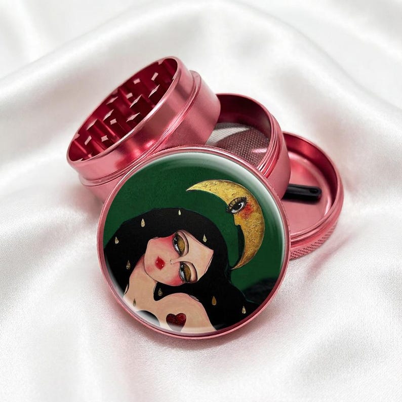 May include: A pink, multi-piece herb grinder with a detailed illustration on the lid. The artwork features a woman with long black hair, a crescent moon, and a green background. The grinder is made of metal.