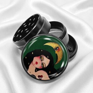 May include: A black metal herb grinder with a decorative top featuring a woman with long black hair, a gold crescent moon, and a green background. The grinder is partially disassembled, revealing the internal components.